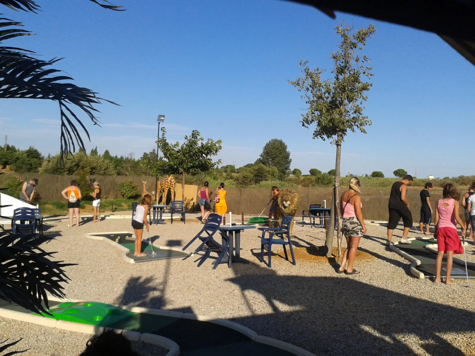 ensemble de personnes jouant au minigolf sur terrain gravillonné et arboré sous ciel bleu